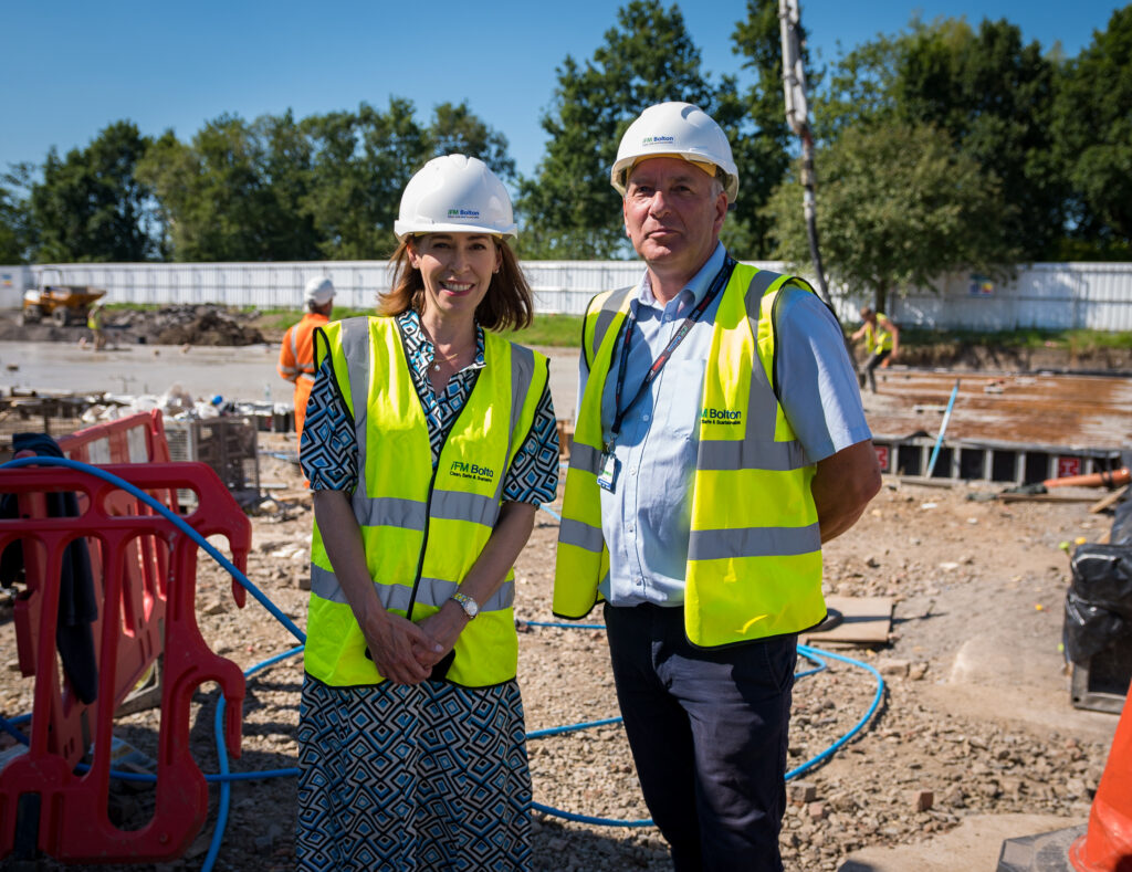 Annette and Cameron stand in front of the concrete slab being poured while wearing