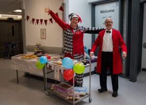 Hayley and Robin stand behind cake sale stall at Royal Bolton Hospital, wearing bright red outfits