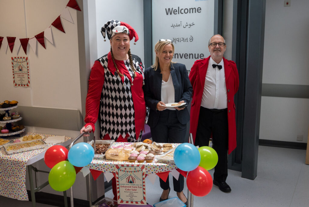 Angela, Hayley and Robin stand behind cake sale stall at Royal Bolton Hospital. Hayley and Robin are wearing bright red outfits, Angela is wearing a suit and is holding a slice of cake. All three are smiling