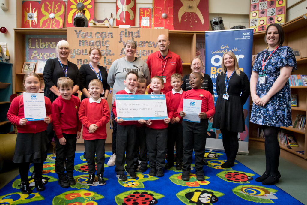 Bolton children charity fundraisers stood in front of Cherry Tree staff and Bolton NHS Foundation Trust staff. Everyone is in a primary school library, smiling at the camera. There are seven adults stood behind eight children