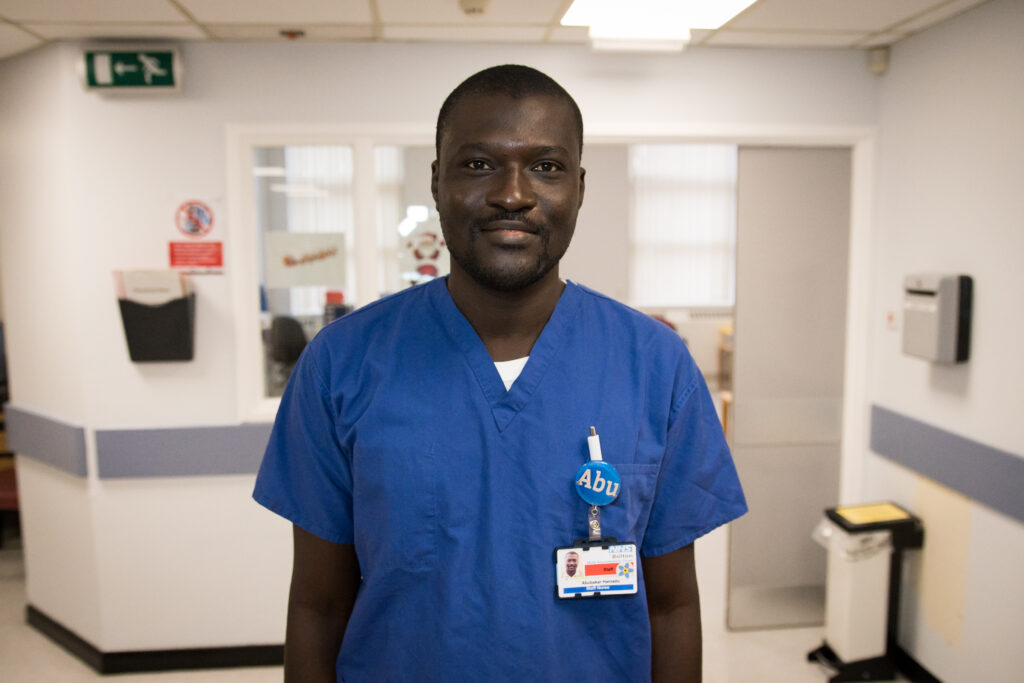 Abubakar Hamadu, an international nurse inspiring others, stands in his blue scrubs in a brightly lit corridor. He is smiling at the camera, and is wearing a bright blue badge saying 'Abu' on it. His hair is short and he has a very closely trimmed goatee.