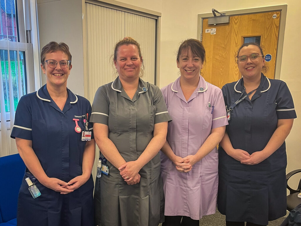 Four ladies stand in a line in an office and are smiling at the camera. All four have dark hair, light skin and are smiling at the camera. They are wearing their uniforms, two in blue, one in grey and one in lilac. They are pictured as Bolton's healthcare staff awarded for dedication and compassion