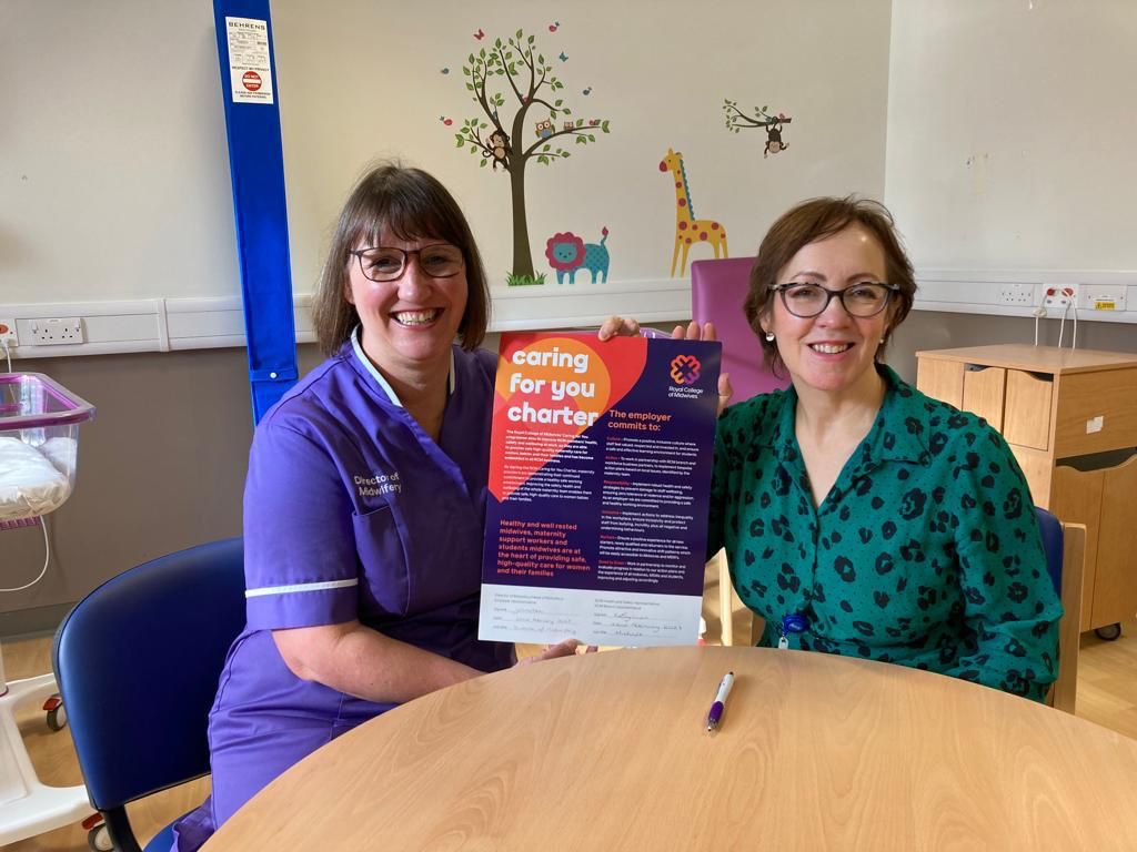 Janet Cotton, Director of Midwifery, holds up a signed Royal College of Midwives Caring For You charter while sat next to Martine, Bolton's RCM rep. They are sat in a post-natal ward room which is light and bright and with cute animal murals on the walls.