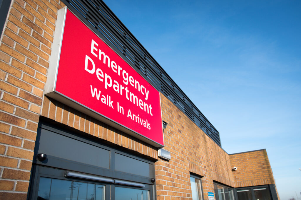 Emergency Department sign at Royal Bolton Hospital. The writing is white, the sign is red. The building is brick and the sky is a lovely shade of blue without a cloud in the sky.