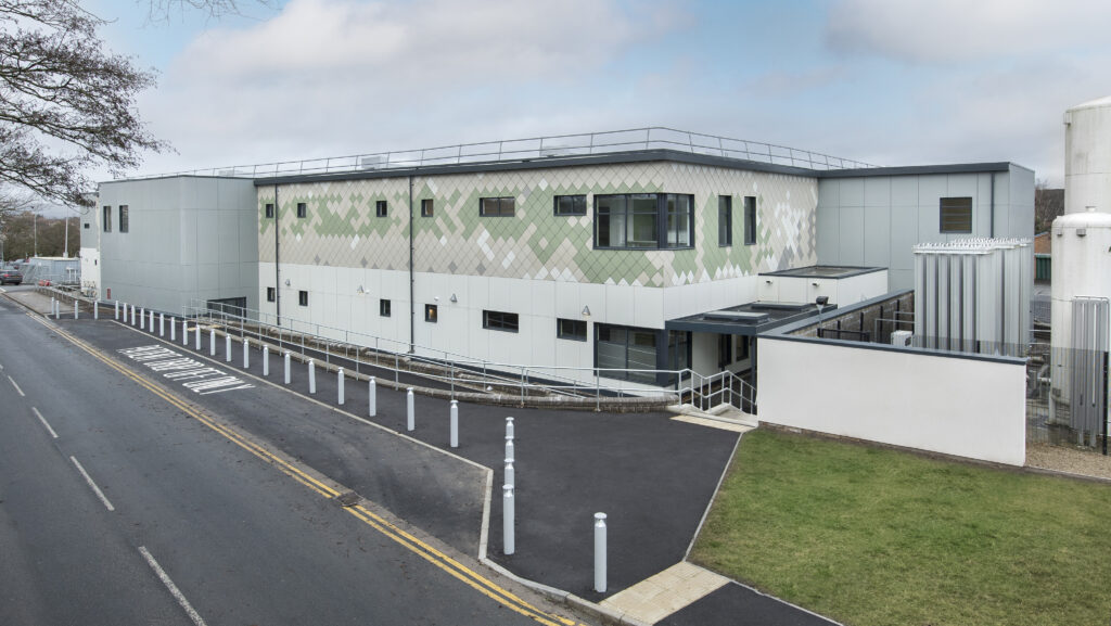 External shot of two-storey theatres building. It is clad in white and green and is quite rectangular in shape. It is next to a road with some trees.