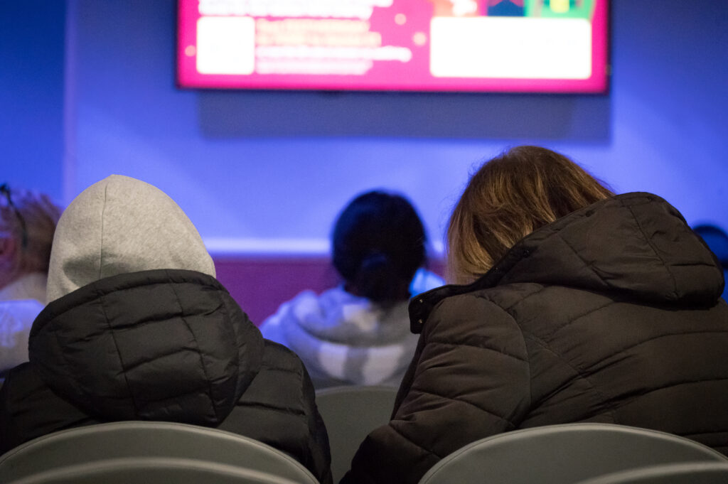 Three people in a waiting room, sat with their backs to the camera. They are looking at a TV on a wall.