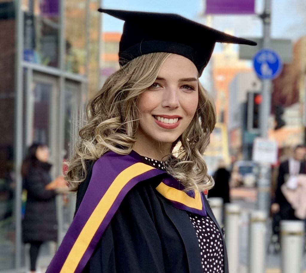 Katie Gibson in her graduation gown outside a university on a sunny day.